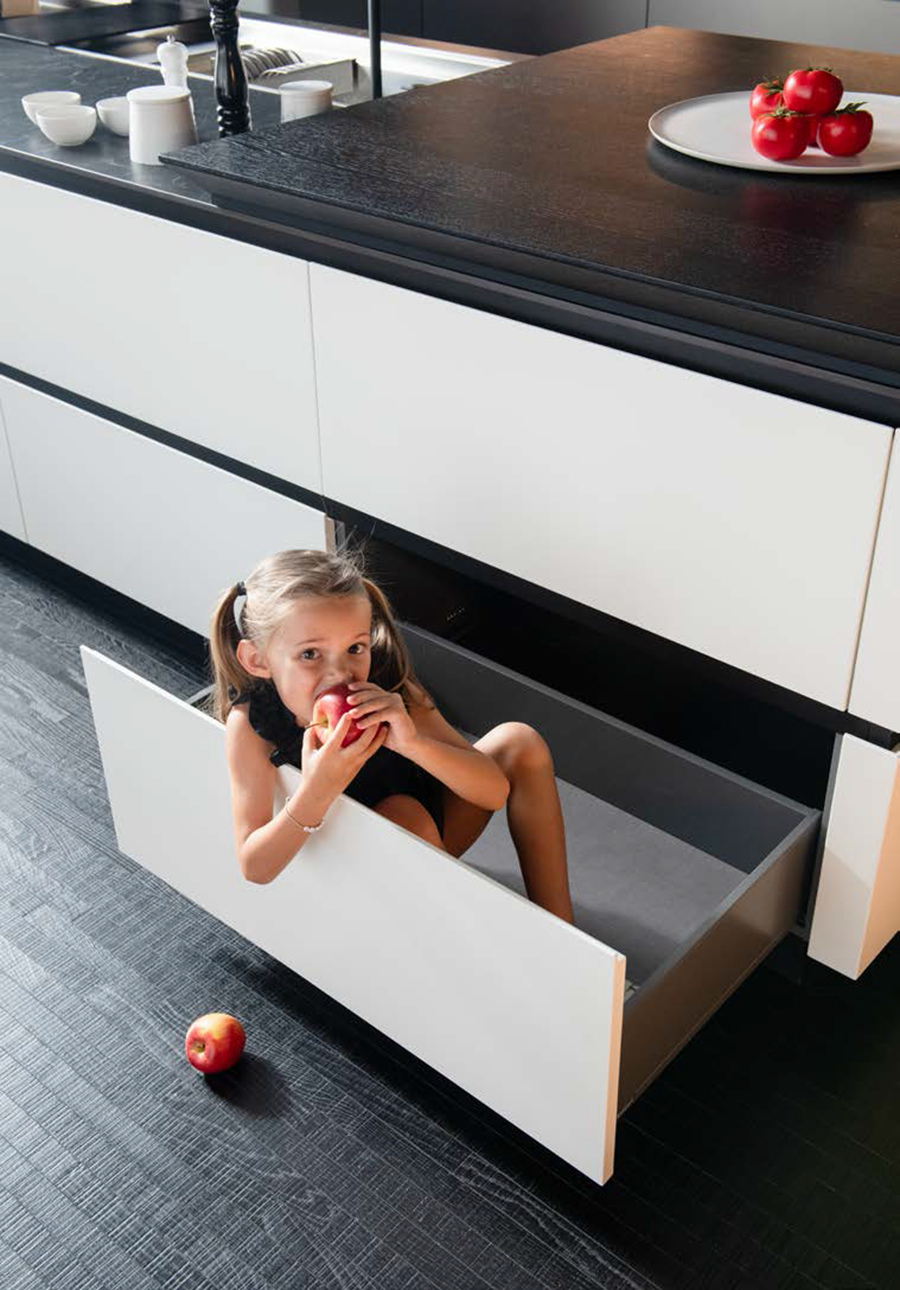 Les matériaux pour une cuisine durable avec CDC Agencement Petite fille avec des couettes, mangeant un pomme rouge dans un meuble de cuisine bas ouvert